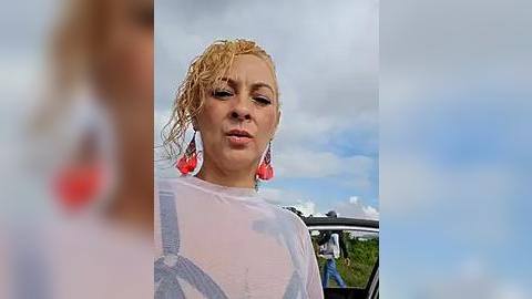 Media: Video of a blonde woman with wet, curly hair, wearing a sheer, wet, white top, and red earrings, standing outdoors with a car in the background under a partly cloudy sky.