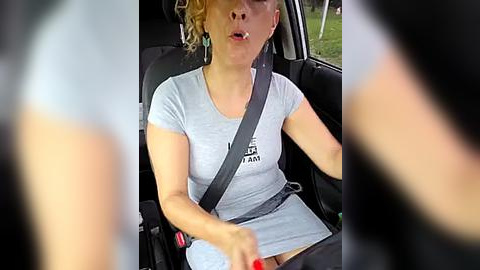 Media: Video of a woman with light skin, short blonde hair, wearing a light gray t-shirt and seatbelt, appearing to be mid-sentence, driving a car with blurred passengers in the background.