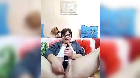 Media: A video of a person with short black hair, glasses, and a blue jacket, sitting on a bed with red and blue pillows, masturbating with a black dildo. Background includes a framed picture and a stuffed dog.