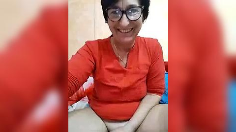 Media: A video of an older woman with short black hair, glasses, and an orange shirt, seated on a bed with red sheets and a blue pillow.