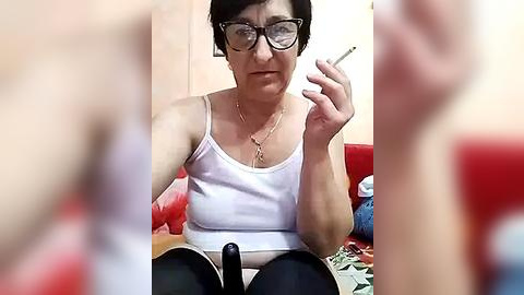 Media: A video of an older woman with short dark hair, glasses, and a white tank top, smoking a cigarette, seated on a red couch in a cluttered room.