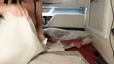 Media: Video of a woman in a white tank top, carrying a white pillow, inside a cramped RV with a messy bed and a red floor.