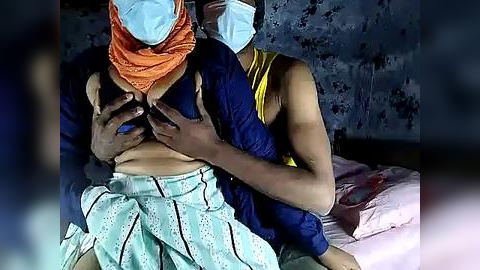 Media: Video of two people, one in an orange headscarf and blue shirt, the other in a yellow tank top and blue shirt, holding each other in a dimly lit, possibly hospital room.