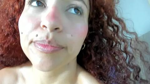 Media: Video of a young woman with light brown skin, curly red hair, and blue eyes, wearing a small nose stud. Her makeup includes a rosy blush and light pink lipstick.