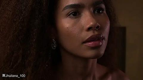 Media: Video of a young Black woman with a medium brown complexion and natural afro hairstyle. She wears minimal makeup, a silver nose ring, and a single earring. The background is blurred, emphasizing her face.