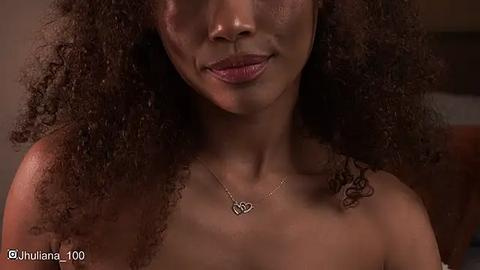 Media: Video of a dark-skinned woman with curly hair and a closed-mouth smile, wearing a heart-shaped necklace, set against a blurred, neutral background.