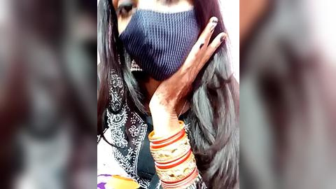 Media: Video of a woman with long black hair, wearing a dark blue mask, a black patterned shirt, and multiple colorful bangles, partially blurred background.