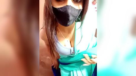 Media: Video of a young woman with long brown hair, wearing a black face mask, light blue tank top, and turquoise skirt, with blurred background.