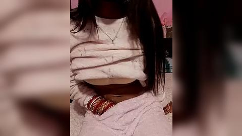 Media: Video of a woman with medium brown skin, wearing a white, long-sleeved sweater lifted to reveal her bare chest, adorned with gold bangles and a necklace, set against a blurred pink background.