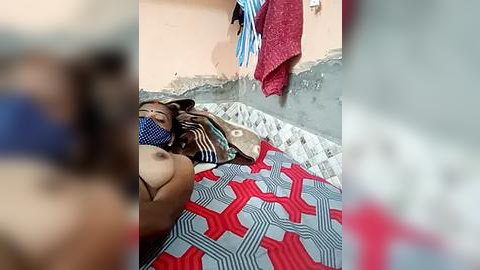 Media: Video of a woman with medium brown skin, wearing a blue and white striped face mask, lying on a red and white patterned blanket in a room with peeling wall paint and a hanging red towel.