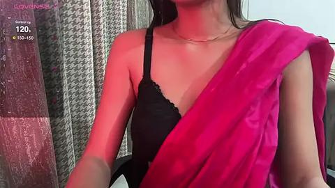 Media: Video of a light-skinned woman in a black lace bra and a vibrant pink saree, her long dark hair partially visible, against a textured curtain background.