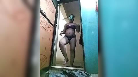 Media: Video of a curvy woman with light skin, wearing a black lace bra and matching panties, standing in a narrow hallway with beige and blue walls.