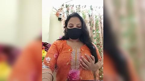 Media: Video of a woman in an orange floral sari with pink roses, black mask, and gold jewelry, holding a phone, in a colorful, festive background.