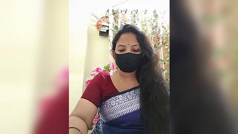 Media: Video of an Indian woman with long black hair, wearing a black mask, red blouse, and blue saree with intricate patterns. She stands in a garden with potted plants and a yellow wall.