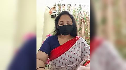 Media: Video of a middle-aged Indian woman with medium skin tone, black hair, and glasses, wearing a black mask, blue blouse, and red sari with white leaf pattern, seated indoors, blurred background.