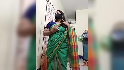 Media: A video of a middle-aged woman in a green saree with a plaid shawl, wearing a black mask, standing in a dimly lit bathroom with a white shower curtain, a fan, and toiletries.