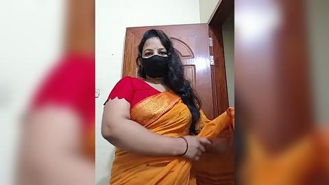 Media: A video of a South Asian woman with medium skin tone, long dark hair, and wearing a red blouse and orange sari, standing in a doorway. She has a black face mask on and holds an orange cloth.