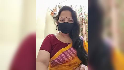 Media: Video of a woman with long black hair, wearing a maroon blouse, yellow and black sari, and black face mask, in a blurred indoor setting with yellow walls and colorful decorations.