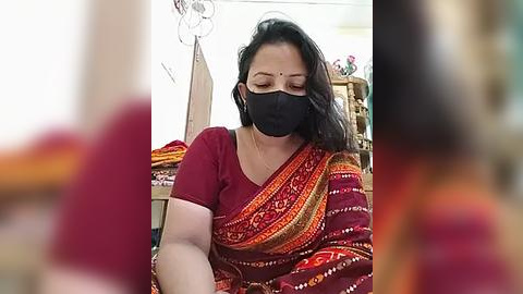 Media: A video of an Indian woman in a red saree with intricate patterns, wearing a black face mask, sewing inside a cluttered room.