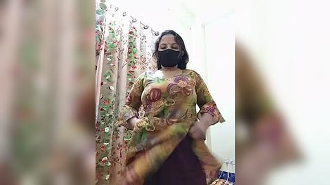 Media: Video of a woman in a floral saree, black mask, and maroon blouse, standing in a brightly lit room with floral curtains.