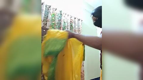 Media: A blurry video of a person wearing a yellow and green saree, standing in a room with floral-patterned curtains and light green walls.