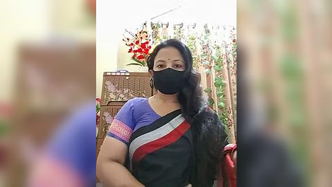 Media: Video of a South Asian woman with medium skin tone and black hair, wearing a black mask and a purple blouse with red and white sash, standing in a garden with red flowers and wooden furniture.