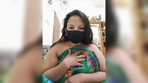 Media: Video of a South Asian woman with medium skin tone, wearing a green sari, black face mask, and holding a colorful cloth over her chest, in a room with decorative objects and a wooden cabinet in the background.