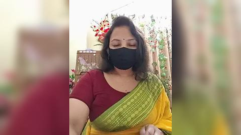 Media: Video of an Indian woman in a maroon top and yellow-green saree, wearing a black mask, sitting indoors with floral decorations in the background.