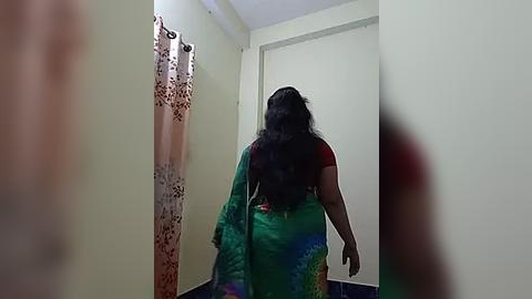 Media: A video of a woman with long black hair, wearing a vibrant green sari with red accents, walking away from the camera in a small, dimly lit room with beige walls and a white door.