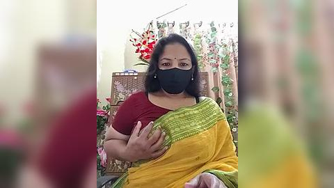 Media: Video of an Indian woman wearing a maroon blouse, black mask, and yellow saree, sitting indoors with a floral backdrop and decorative plants.