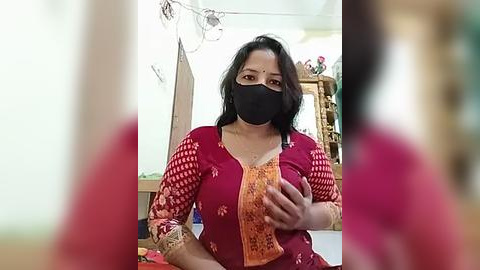 Media: Video of a South Asian woman with medium skin tone, wearing a red kurta with intricate embroidery, black mask, and black hair, holding her chest, indoors with blurred background.