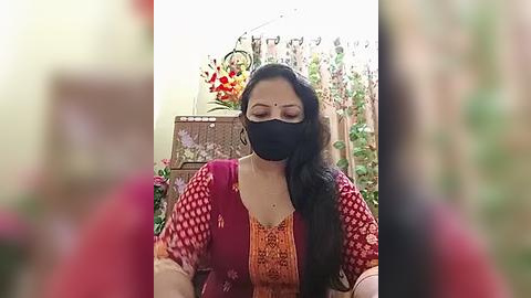 Media: Video of a South Asian woman with long black hair, wearing a red and orange traditional dress, black face mask, and a red bindi, seated indoors with floral decor in the background.