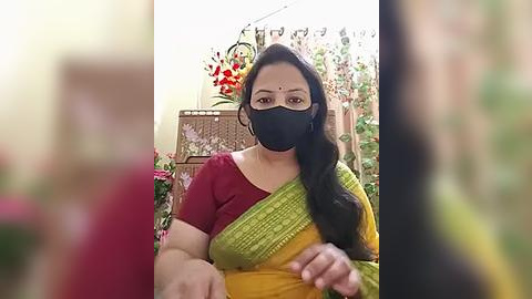 Media: Video of an Indian woman in a yellow-green sari, black face mask, and red blouse, sitting on a wicker chair with flowers in the background.