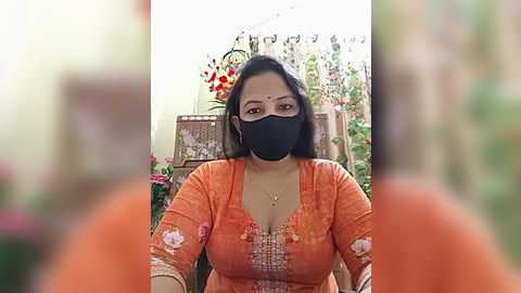 Media: Video of a woman in a floral orange saree, black mask, and glasses, seated indoors amidst vibrant floral decorations.