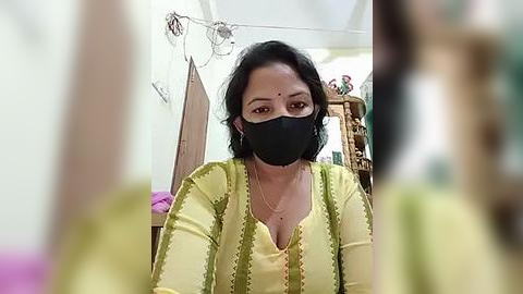 Media: Video of a South Asian woman with medium skin tone and short black hair, wearing a yellow kurta with green embroidery, black mask, and a nose stud, in a dimly lit room with Hindu religious idols and a wooden door.