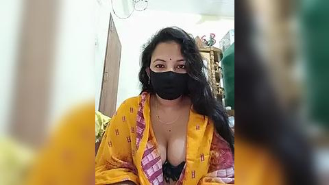 Media: Video of a South Asian woman with long black hair, wearing a yellow saree with pink and purple patterns, a black face mask, and a black bra visible under the saree. The background shows a cluttered room with household items.