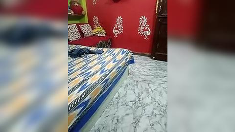 Media: Video of a colorful bedroom with a bed covered in a striped blue, yellow, and white bedspread. Red walls feature white, stylized bird patterns. A cluttered wooden dresser is in the background.