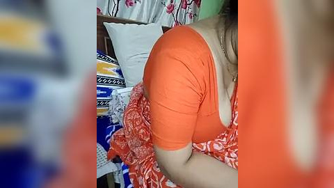 Media: A video of a woman in an orange top and patterned orange and white skirt, lying on a bed with colorful bedding, including a blue and white patterned pillow.