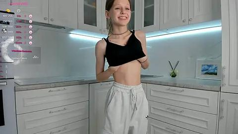 Media: Video of a smiling young woman with light skin and brown hair, wearing a black crop top and white sweatpants, standing in a modern kitchen with white cabinets and a blue LED strip.