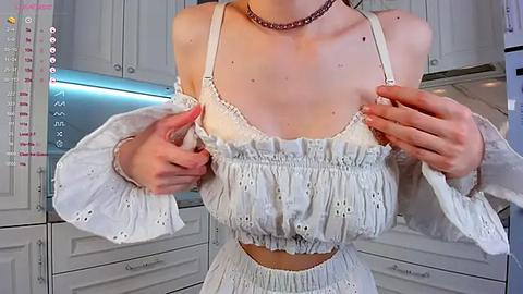 Media: Video of a light-skinned woman in a white off-shoulder top and high-waisted skirt, adjusting her top straps. She wears a pink choker and is in a modern, white kitchen with stainless steel appliances.