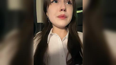 Media: A video of a young woman with long brown hair, fair skin, and blue eyes, wearing a white blouse, looking contemplative, blurred figures in the foreground.