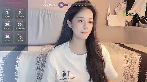 Media: Video of a young East Asian woman with long black hair, wearing a white t-shirt, sitting on a bed with a black and white striped blanket. The room has a modern, minimalistic decor.