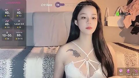 Media: Video of a young Asian woman with long black hair, fair skin, and a tattoo on her shoulder, wearing a white lace lingerie top, lying on a bed with a striped blanket.