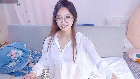 Media: Video of a smiling East Asian woman with long straight hair, wearing glasses, a white blouse, and a blue blanket, sitting on a bed with a gray headboard and plush toys.