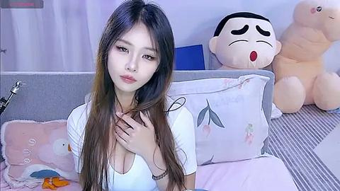 Media: A video of an Asian woman with long brown hair, fair skin, and medium-sized breasts in a white top, sitting on a bed with plush toys, including a Pac-Man plush and a teddy bear, in a soft, pastel-themed room.