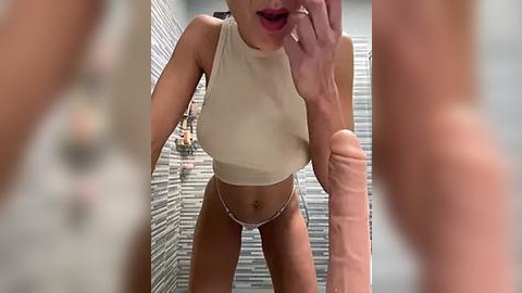 Media: Video of a slender, tan-skinned woman with large breasts wearing a sheer, beige crop top and white thong, standing in a tiled shower.