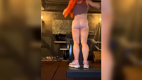 Media: A video captures a woman with long orange hair, wearing a grey sports bra and white shorts, standing on a weight bench in a dimly lit gym, with a blurred figure in the foreground.