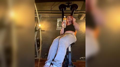 Media: Video of a young woman with red hair, wearing a black tank top and light blue sweatpants, sitting on a weight bench in a dimly lit, industrial-style gym, holding a water bottle.