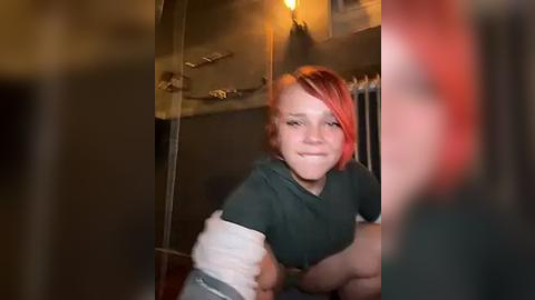 Media: A video of a young woman with short, bright red hair and fair skin, wearing a dark green shirt, posing in a dimly lit, rustic setting with exposed wood beams and a metal shelf.