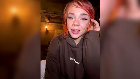 Media: Video of a young woman with dyed pink hair, wearing a black hoodie, smiling, sitting indoors with dim lighting, blurred background.